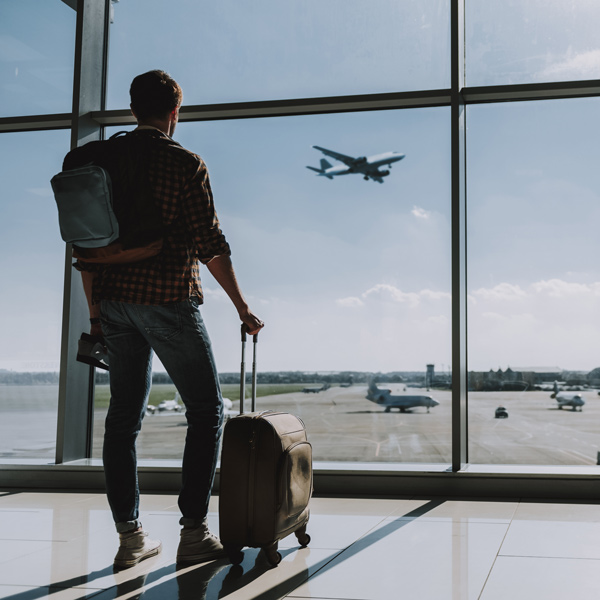 man in airport with rucksack and suitcase looking out of glass windows at plane taking off